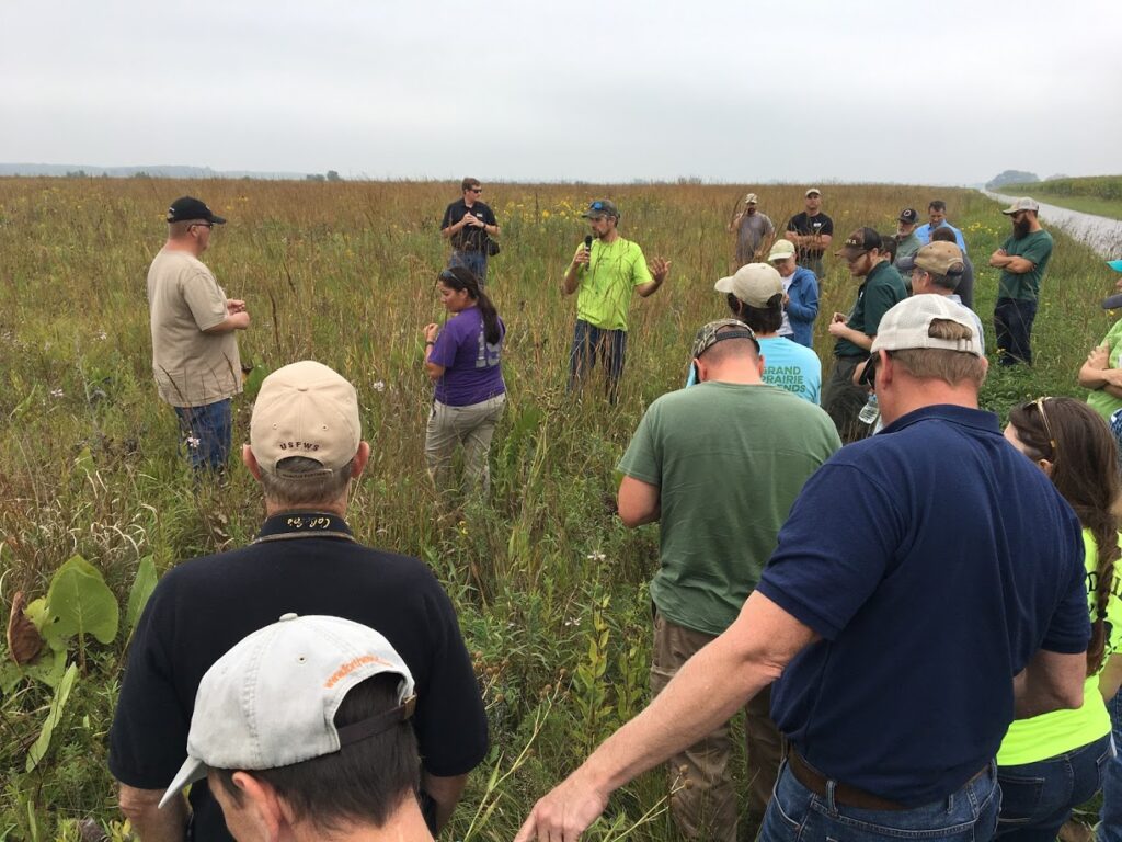 Group of people in tall grass with one man in the middle holding a mic.